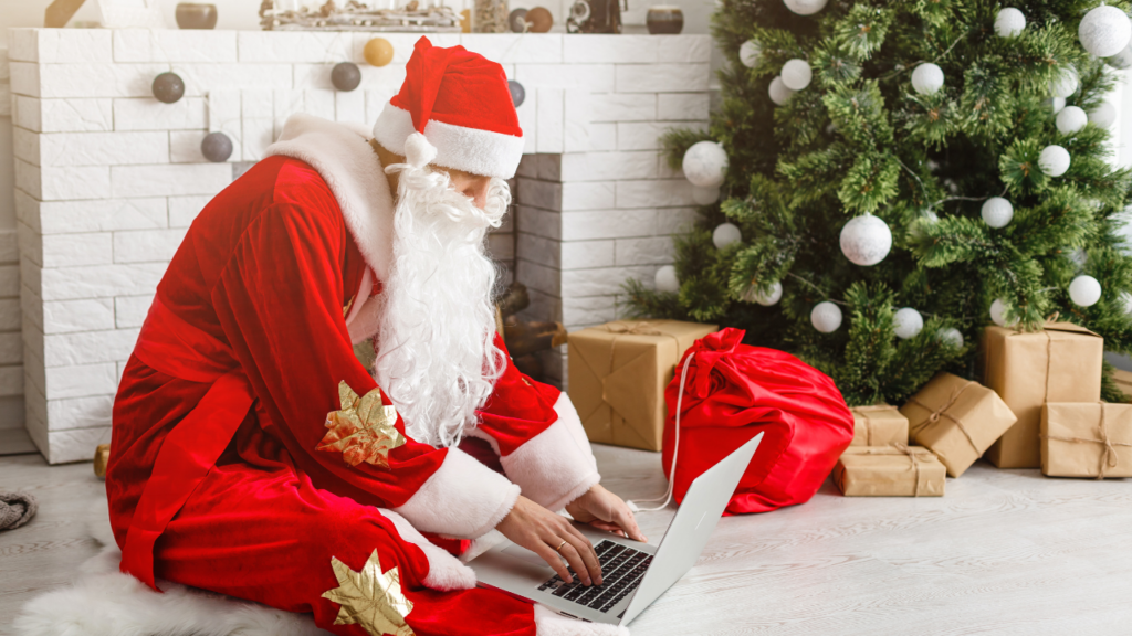 Santa shopping online, sitting on the floor in front of a Christmas tree with packages around it.